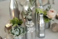 a chic and simple wedding centerpiece made of silver bottles and jars, with greenery, blooms and succulents plus candles around