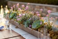 a long wooden planter with succulents and small pink blooms over a striped table runner is a cool backyard centerpiece idea