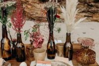 a lovely and simple fall boho cluster wedding centerpiece of beer bottles, with pampas grass, bold blooms, greenery and wildflowers plus candleholders around