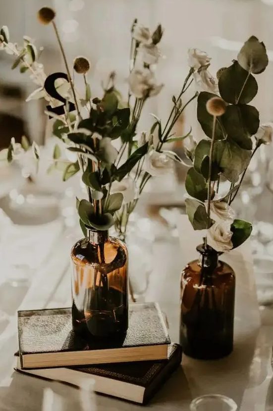 a lovely cluster wedding centerpiece of a stack of books, apothecary bottles with neutral blooms and greenery is a cool idea