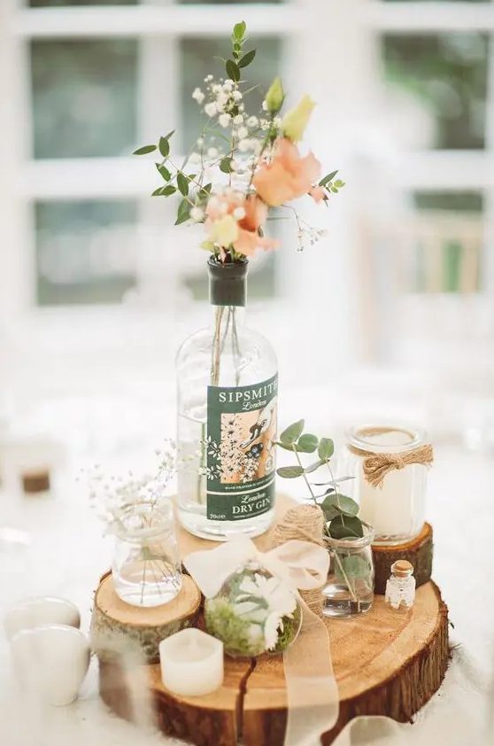 a quirky rustic wedding centerpiece of tree and branch slices, bottles and jars with pink and white blooms, greenery, moss and candles