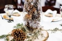 a simple rustic winter wedding centerpiece of a wood slice, a tall glass with pinecones and evergreens is veyr easy and fast to DIY