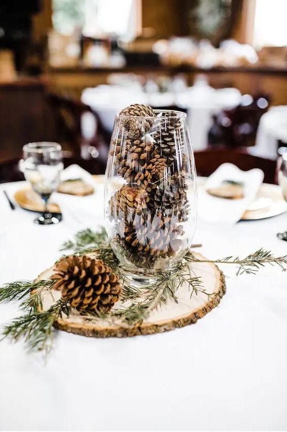 a simple rustic winter wedding centerpiece of a wood slice, a tall glass with pinecones and evergreens is veyr easy and fast to DIY