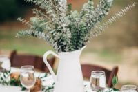 a white milk jug with eucalyptus, a greenery table runner and dark candle holders with candles