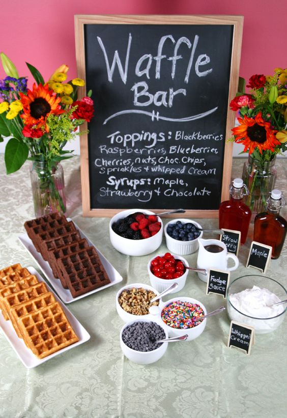 a mini waffle bar with a chalkboard sign, waffles, toppings and berries and some syrups is cool