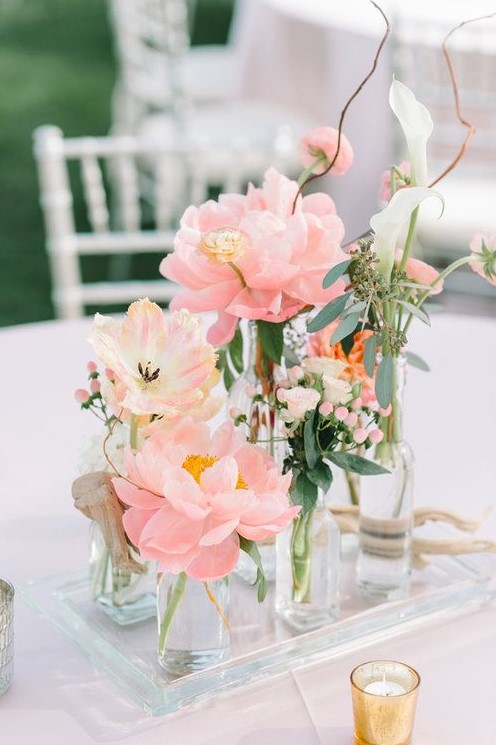 a beautiful cluster wedding centerpiece of clear bottles, blush peonies, white callas, greenery and twigs is a chic idea for spring or summer