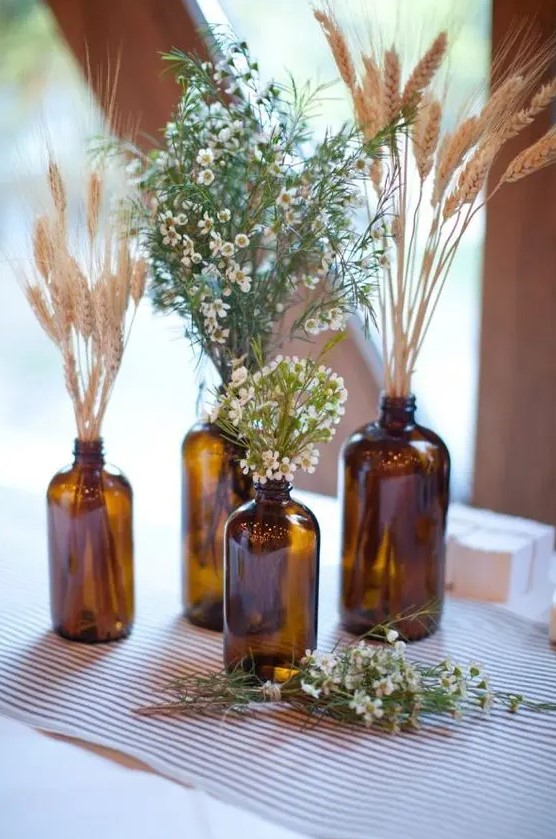 a boho cluster wedding centerpiece of apothecary bottles, wheat, greenery and waxflower is a cool decoration to DIY