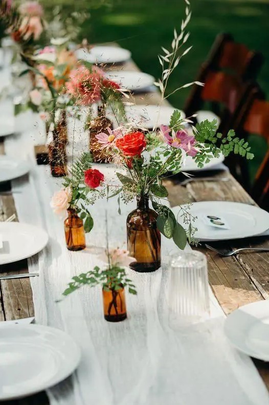 a bright apothecary bottle cluster wedding centerpiece with pink and red blooms, greenery and simple grasses for a relaxed and rustic summer wedding