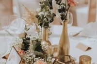 a cluster fall wedding centerpiece of gilded bottles and candleholders, greenery, white and pink roses, candles and baby’s breath