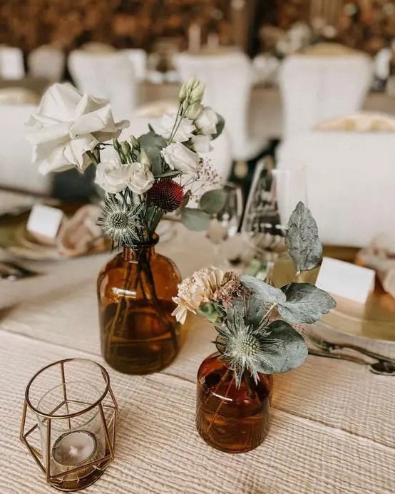 a cluster wedding centerpiece of apothecary bottles, greenery and thistles, neutral blooms and candles around is a cool idea for a boho wedding