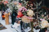 a cluster wedding centerpiece of dark bottles with bright and pastel blooms, with greenery and berries plus candles
