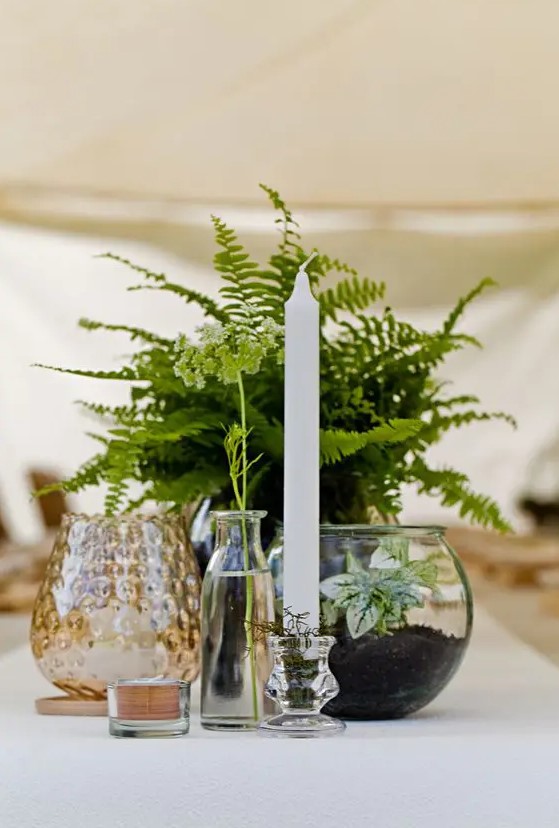 a cluster wedding centerpiece of potted fern, a candle, some candleholders and a terrarium with greenery