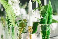 a cluster wedding centerpiece of vintage glasses, fern, greenery, white blooms and a candle in a gold candleholder