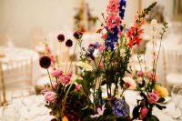 a colorful boho cluster wedding centerpiece of bold blooms and greenery in bottles and vases, petals on the table and a cool table number