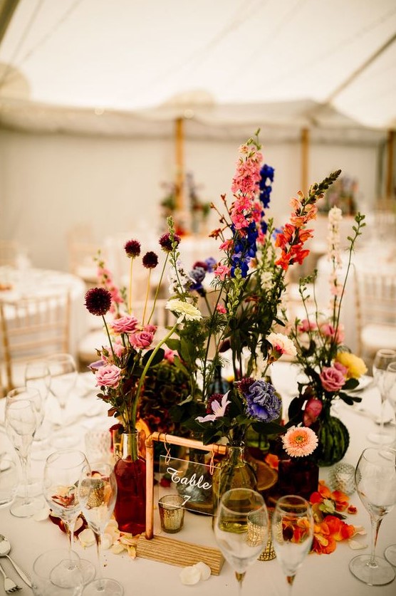 a colorful boho cluster wedding centerpiece of bold blooms and greenery in bottles and vases, petals on the table and a cool table number