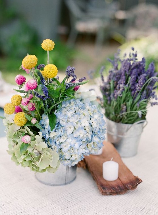 a colorful rustic cluster wedding centerpiece of buckets with hydrangeas, billy balls and lavender plus a wooden piece with a candle