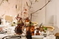 a fall cluster wedding centerpiece of apothecary bottles, blooms and bold foliage, branches and greenery is a cool idea