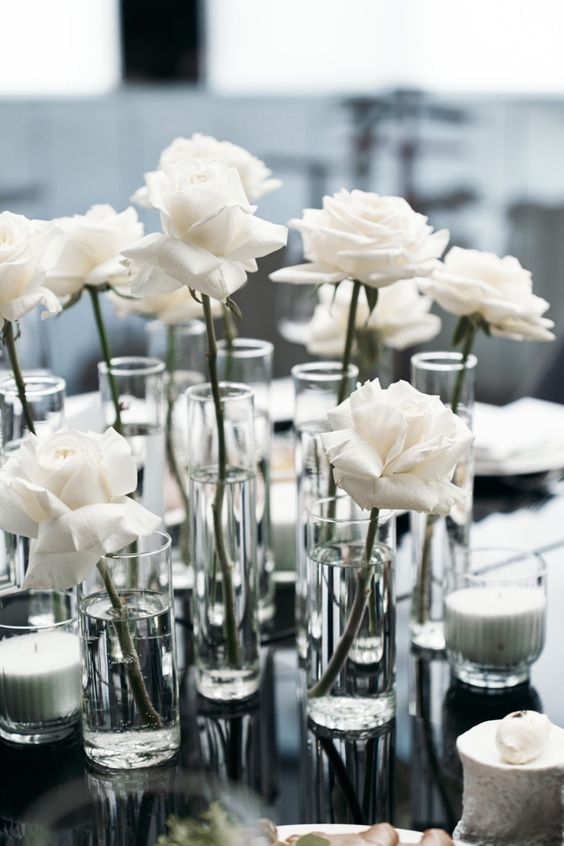 a minimalist wedding centerpiece of glasses with single white roses and pillar candles is a cool and chic idea