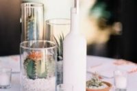 a modern cluster wedding centerpiece of a white bottle with a table number, glasses with cacti and succulents and mini candles around