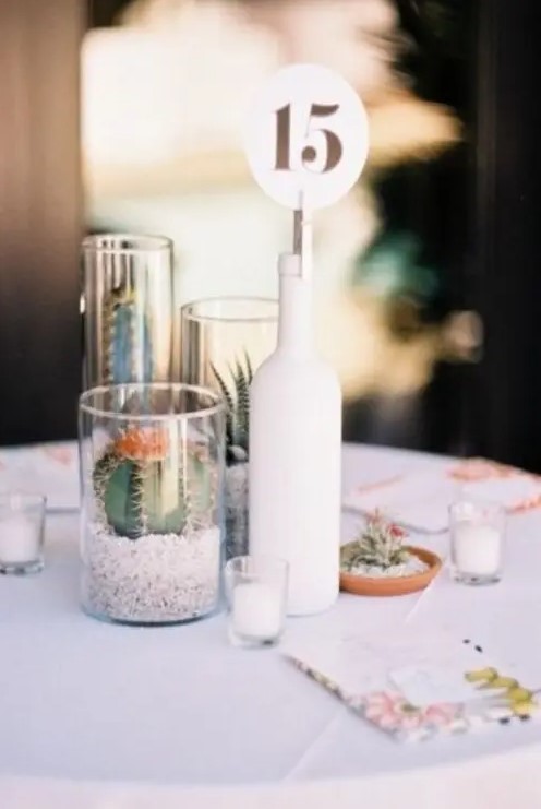 a modern cluster wedding centerpiece of a white bottle with a table number, glasses with cacti and succulents and mini candles around