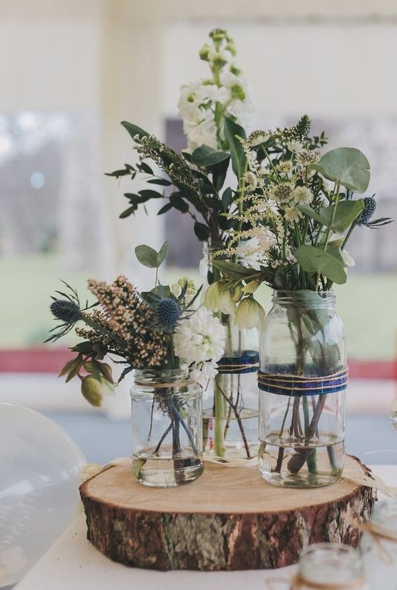 a rustic cluster wedding centerpiece of a wood slice, jars, neutral and blush blooms, greenery and thistles is wow