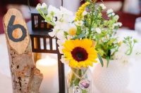a rustic cluster wedding centerpiece with sunflowers, white blooms, a candle lantern and a tree branch with a table number