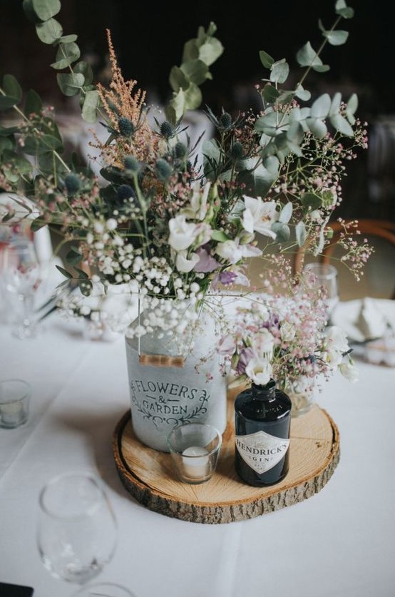 a rustic wedding centerpiece of a wood slice, bottles with white blooms, thistles and greenery and a single candle is wow