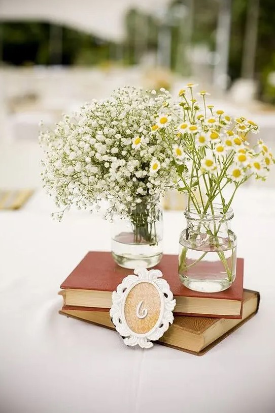 a vintage rustic wedding centerpiece of a stack of books and two vases, one with baby's breath and the second with daisies