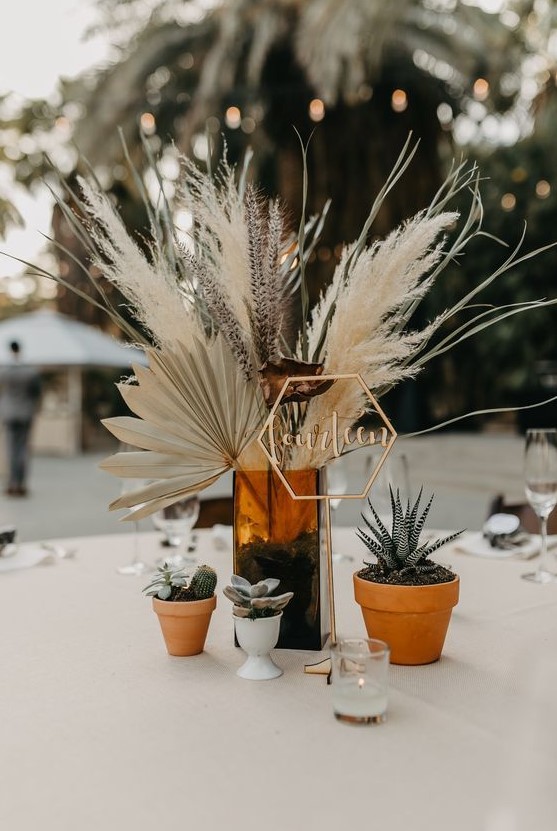 a wedding centerpiece of an amber glass vase, pampas grass, lavender, dried fronds and some potted succulents around
