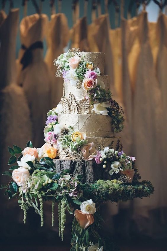 a bark wedding cake topped with pastel blooms and greenery and served on a stand with moss, foliage and blooms