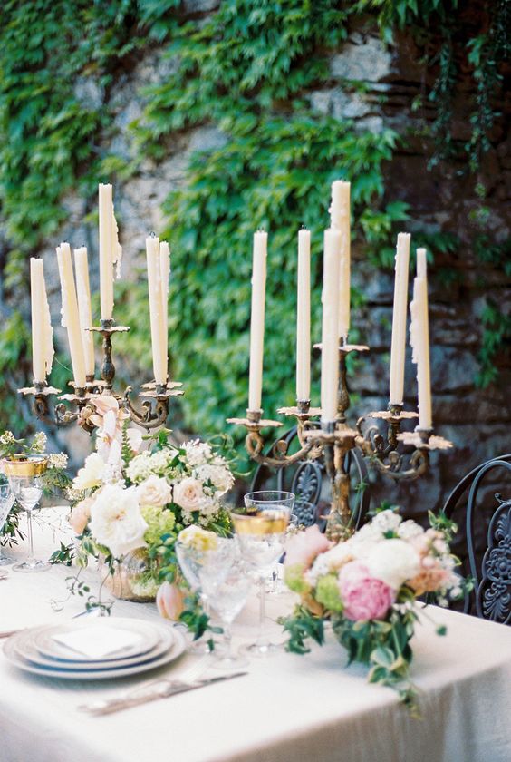 a romantic fairy tale wedding tablescape with blush and white blooms, greenery, candles in vintage candleholders and elegant glasses