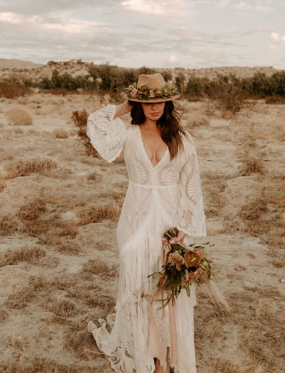a boho lace A line wedding dress with a plunging neckline, bell sleeves and long fringe plus a train and a flower detailed hat