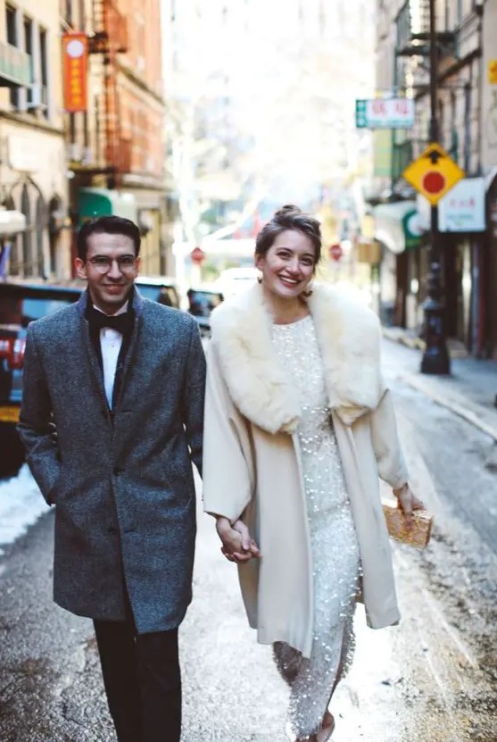 a glam winter bride wearing an embellished wedding dress, a beautiful white coat with a fur collar and rocking a white and gold clutch