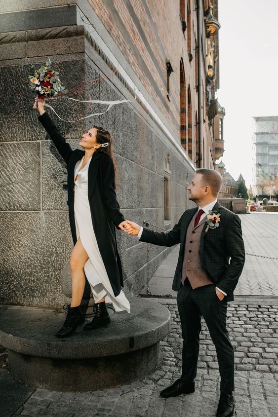 a neutral wedding dress, black boots and a black coat are a cool and bold look for a winter bride