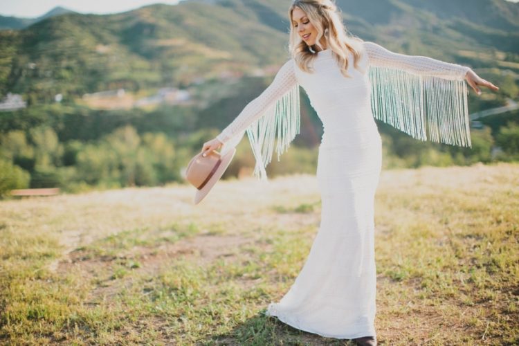 a textural sheath wedding dress with long sleeves detailed with long fringe and a hat for a boho bridal look