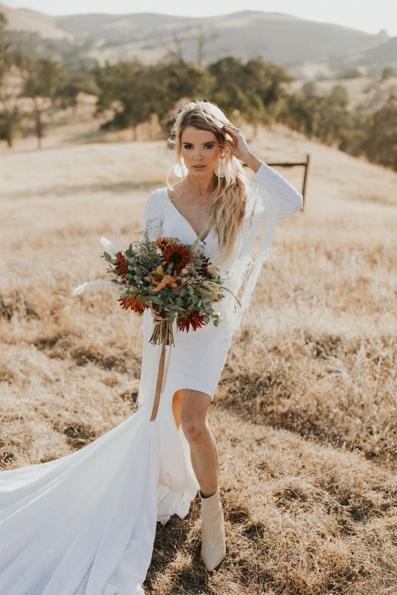 a white plain wedding dress with a high low skirt with a train, a V-neckline, long sleeves with fringe for a boho bride