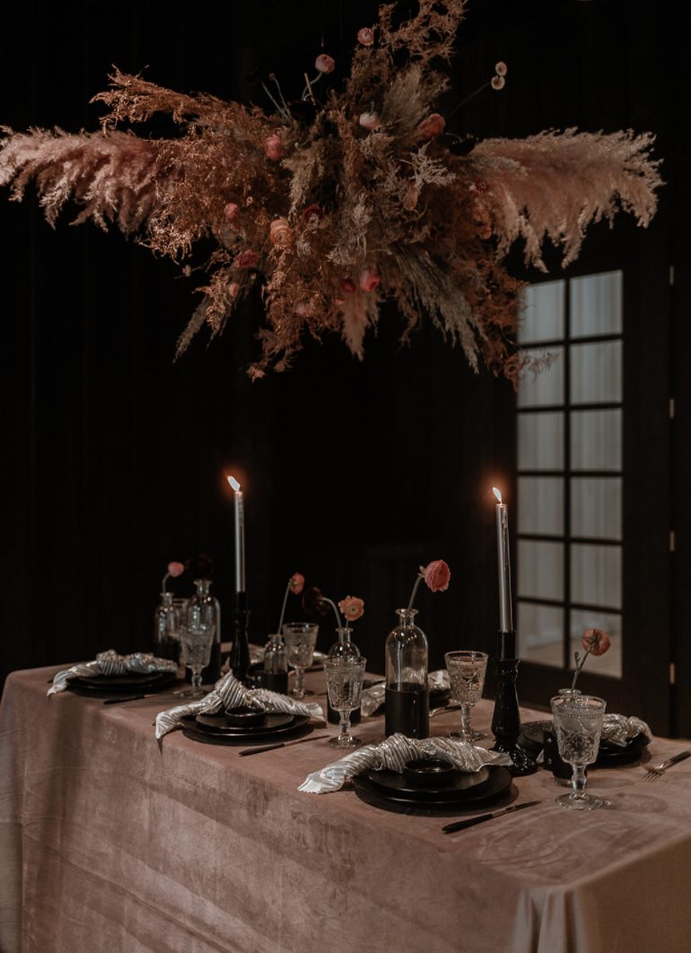 The wedding tablescape was done with a terracotta tablecloth, a dried grass and bloom overhead hanging, black candleholders and plates and silver napkins
