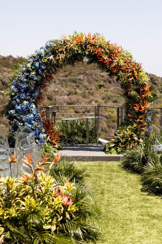 a gradient floral and greenery wedding arch from purple and blue to yellow, rust and orange finished with tropical leaves