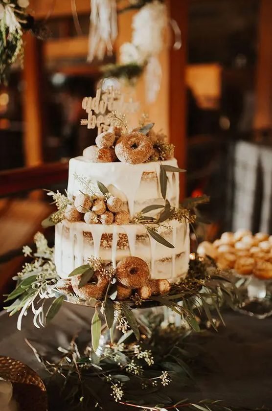 a boho naked wedding cake with creamy drip, greenery, donuts and candies plus a calligraphy topper