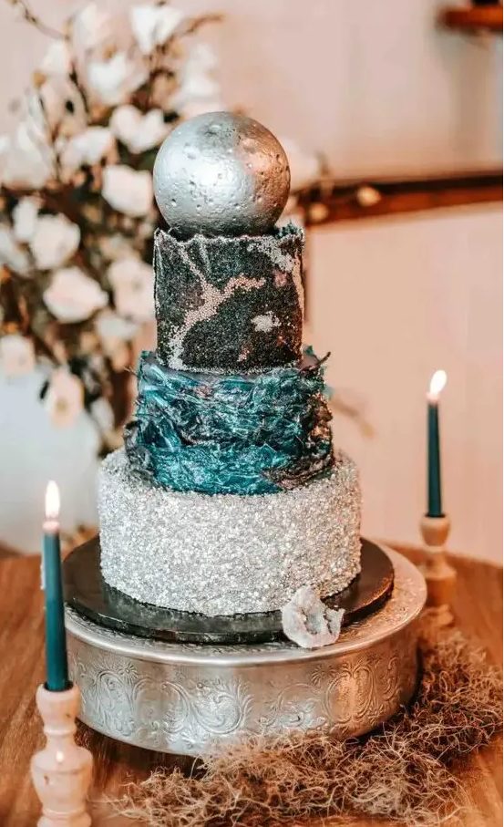 a celestial wedding cake with a silver tier, a teal textural one, a black tier with silver decor and a silver sphere on top
