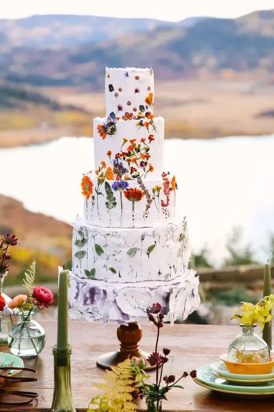 A multi tier white wedding cake with pressed bold blooms, a tier with only leaves and a painted lilac tier is amazing for summer