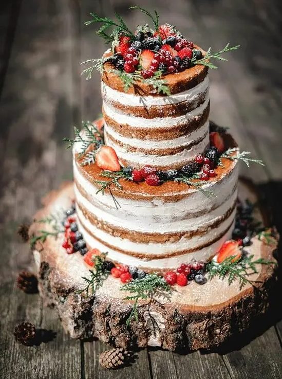a naked wedding cake topped with sugared berries, greenery served on a wood slice for a rustic wedding
