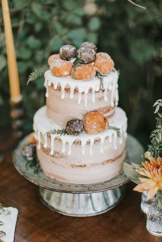 a naked wedding cake with creamy drip, donuts, greenery and fern is a cool wedding dessert for a rustic wedding