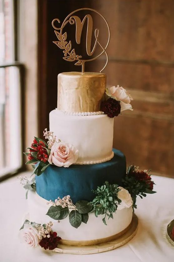 a stylish wedding cake with a gold, two white and teal tiers, white, blush and burgundy blooms and greenery plus a calligraphy topper
