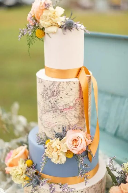 A vintage inspired wedding cake with a blue and white tier, with a map tier and natural blooms and marigold ribbons