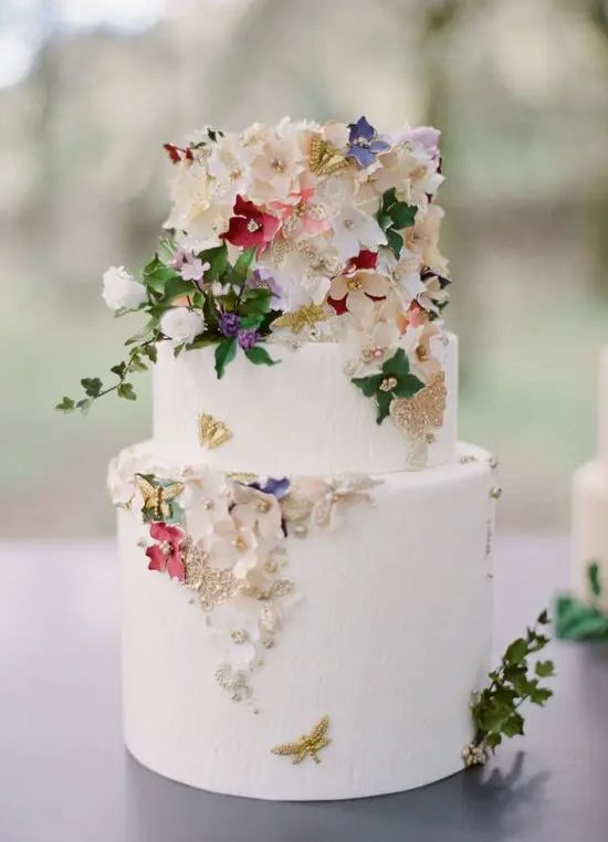 a white buttercream wedding cake decorated with gold foil, sugar flowers and real leaves, beads and butterflies is amazing
