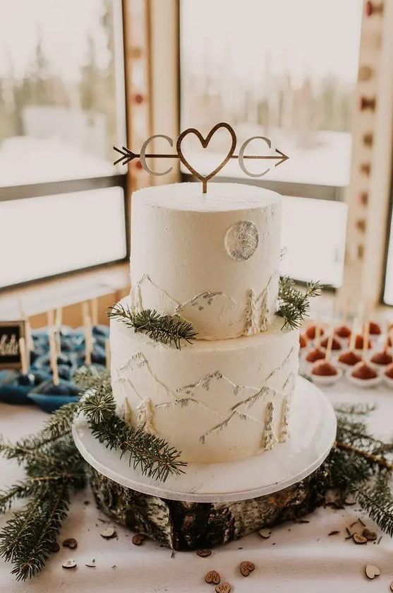 a white buttercream wedding cake with painted mountains, a moon and trees, with evergreens and a monogram and heart cake topper