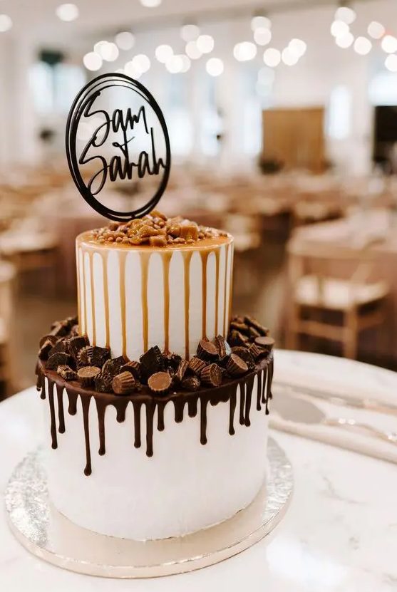 a white wedding cake with caramel and chocolate drip, with candies on top and a modern cake topper is wow