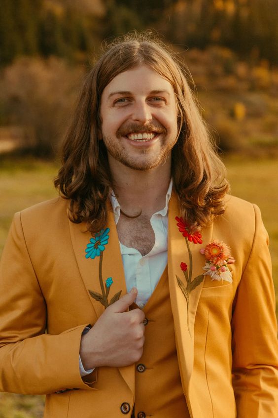 a bold boho groom's look with a mustard three-piece suit, a white shirt, bold floral embroidery and a dried flower boutonniere