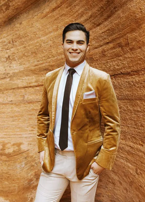 a catchy groom's outfit with a white shirt, neutral pants, a black tie, a mustard velvet blazer for the fall